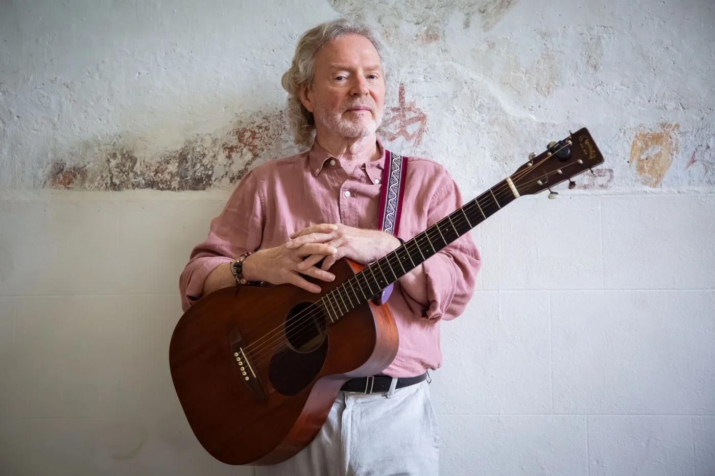 Reg Meuross holding an acoustic guitar against a whitewashed wall